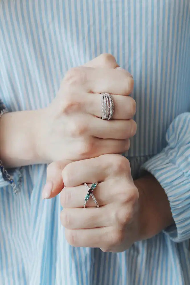Two hands wearing rings, one with multiple thin bands, the other with a criss-cross design featuring dark and light blue stones.